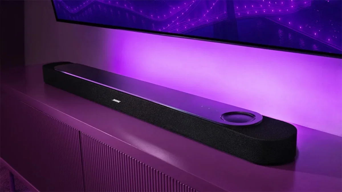 Bose Lifestyle Ultra Soundbar en negro sobre mueble de listones, con iluminación púrpura y TV en la pared; se ve la cara superior de vidrio y el logo Bose.