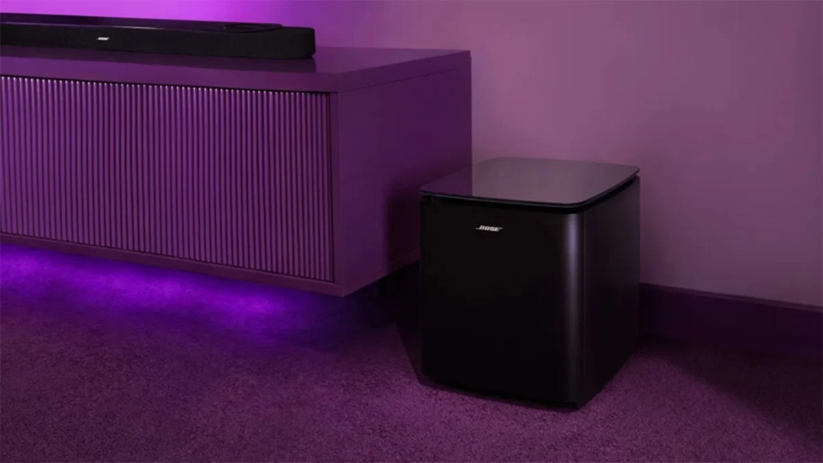 Bose Lifestyle Ultra Subwoofer cúbico en negro con tapa de vidrio, sobre alfombra junto al mueble; arriba asoma la soundbar, en ambiente con luz púrpura.