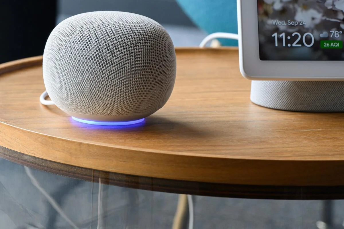 Google Home Speaker Porcelain con el anillo de luz encendido sobre una mesa de madera, al lado de un Nest Hub