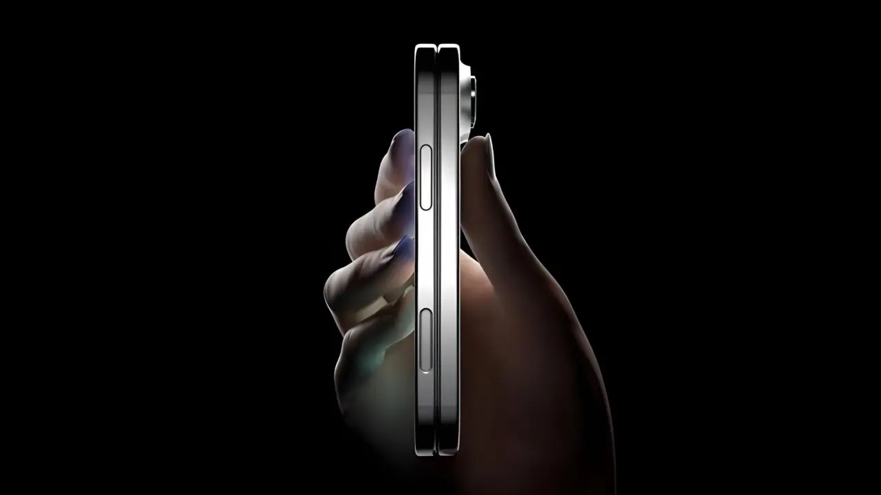 Mano sosteniendo un iPhone plegable cerrado visto desde el perfil sobre un fondo negro. Resalta su extrema delgadez, el acabado metálico brillante de los bordes y los botones laterales.
