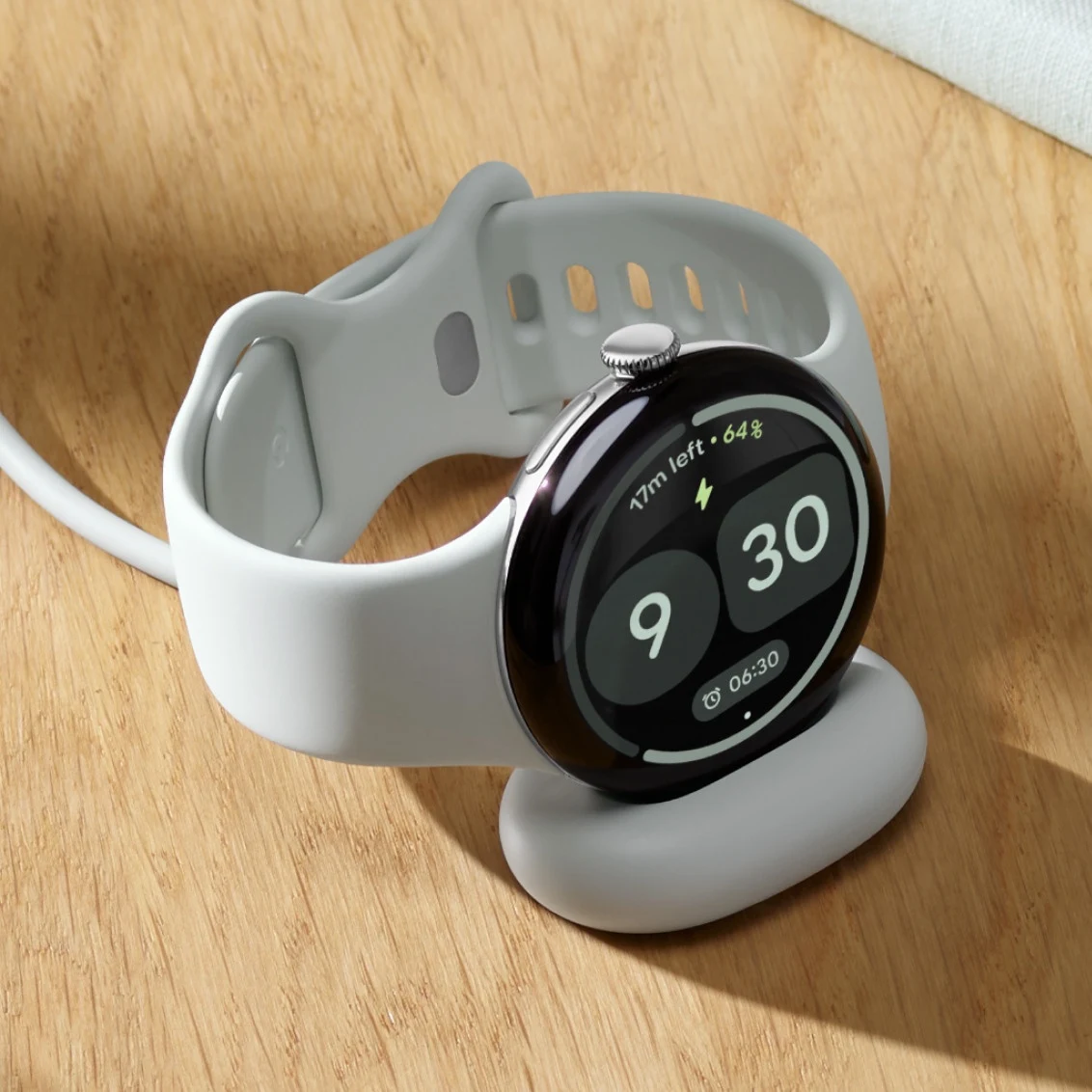 El Pixel Watch 5 apoyado en su base de carga sobre una mesa de madera mostrando el tiempo restante